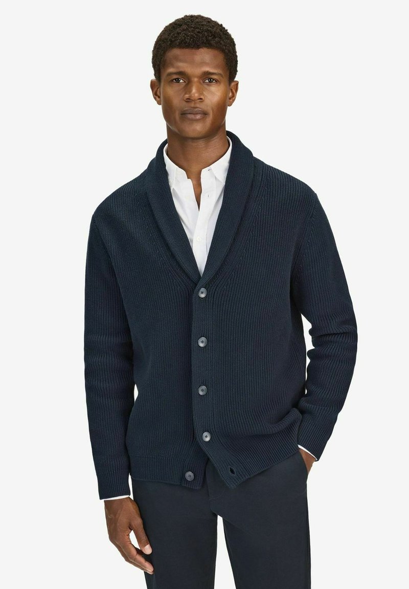 Mörk marinblå kavaj med ribbad textur, sjalkrage och fem knappar, bärs över en vit skjorta och matchas med mörka byxor.