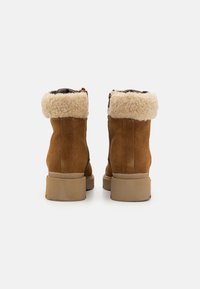 Botas de tobillo de ante marrón con un puño forrado de piel de oveja, suelas planas de goma beige y una cremallera lateral para poner y quitar fácilmente.