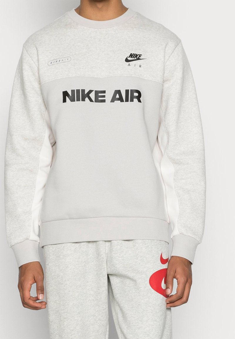 Ljusgrå Nike-tröja med "NIKE AIR" i fet svart text, utrustad med rund halsringning och kontrasterande vita sidopaneler.