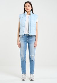 Šviesiai mėlynas be rankovių pūkinis liemeniukas su užtrauktuku, derinamas su baltu marškinėliu ir nublukusiais šviesiai mėlynais džinsais. Balti sportbačiai užbaigia įvaizdį.