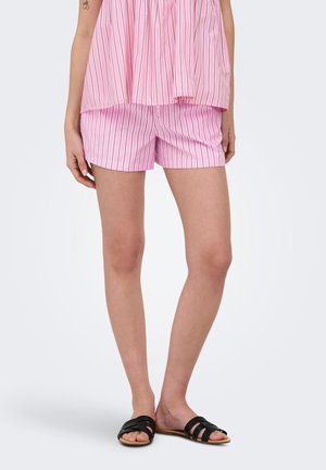 Vrouw draagt roze-wit verticale gestreepte short en bijpassende losse top, met zwarte slip-on sandalen, staand tegen een witte achtergrond.
