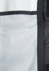 Tela de malla negra con un patrón texturizado, asegurada con un borde de tela negra y un cierre de velcro. El fondo es de un material suave y de color claro.