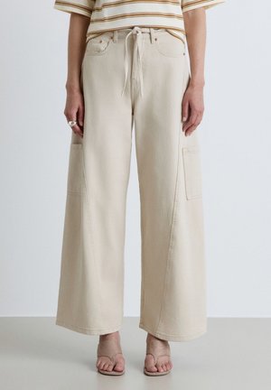 Personne portant un pantalon beige à jambes larges avec poches latérales, un haut rayé et des sandales beige ouvertes à l'avant, debout devant un fond uni.