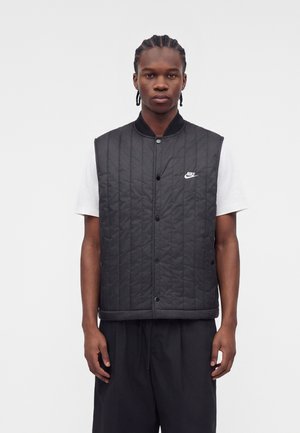 Jeune homme aux cheveux tressés portant un gilet matelassé noir Nike par-dessus un t-shirt blanc et un pantalon noir, debout devant un fond uni.