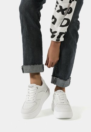 Sneakers bianchi con lacci, tomaia perforata, suola elevata e texture in pelle liscia, abbinate a jeans scuri in denim arrotolati.
