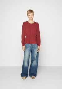 Blouse bordeaux à manches longues avec taille froncée, associée à un jean bleu délavé à jambes larges. L'ensemble présente une coupe décontractée et une texture douce.