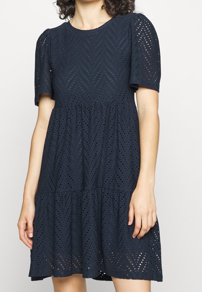 Femme portant une robe bleu marine en œillet avec des manches courtes bouffantes et une jupe à étages, avec des motifs découpés en forme de chevrons.