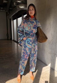 Γυναίκα που στέκεται σε εσωτερικό χώρο, φορώντας ένα σετ με παλτό και παντελόνι με σχέδιο πεϊζλί, πορτοκαλί μαντήλι στο λαιμό και κρατώντας μια μεγάλη καφέ σουέντ τσάντα.