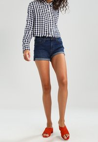 Chemise à carreaux bleu marine et blanc, short en jean foncé et mules rouges à bout ouvert. La chemise est dotée de boutons et de manches longues.