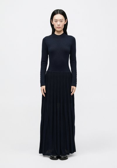 Navy long-sleeved knit dress with a fitted bodice and pleated skirt. Features button accents on sleeves and smooth texture. Worn with chunky shoes.