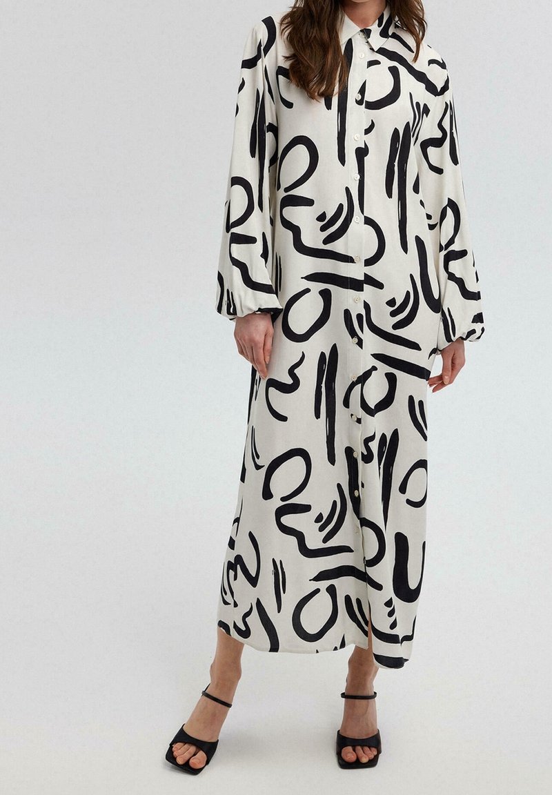 Femme portant une longue robe blanche boutonnée avec des motifs abstraits noirs en coups de pinceau et des talons ouverts noirs, debout devant un fond uni.