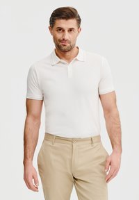 Biała koszulka polo z kołnierzykiem i krótkimi rękawami, z kontrastowym wykończeniem. W połączeniu z beżowymi spodniami, prezentującymi czysty, casualowy wygląd.