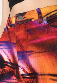 Jeans de ganga multicoloridos com padrões vibrantes em laranja, roxo e rosa, apresentando um botão de metal e costuras contrastantes na cinta.