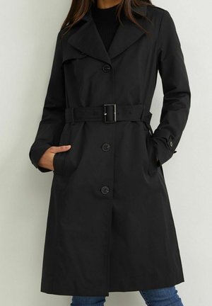 Sort trenchcoat med bælte og knaplukning, båret over sort top og blå jeans, hænderne i lommerne, foran en ensfarvet lys baggrund.