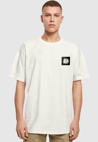 Hvid bomulds t-shirt med en afslappet pasform, med et sort firkantet lomme design og en hvid tusindfryd grafisk på venstre brystområde.
