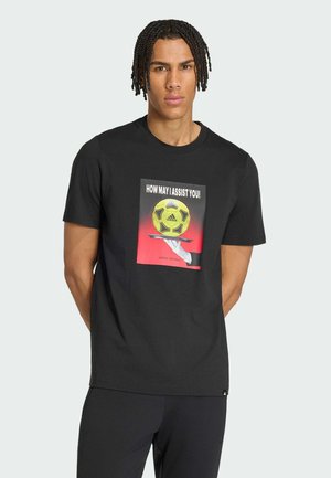 Homme aux cheveux tressés portant un t-shirt noir avec un graphisme d'une main tenant un plateau avec un ballon de football Adidas jaune et le texte « Comment puis-je vous aider ! »