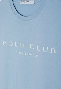 Světle modré tričko z organické bavlny s bílým potiskem "POLO CLUB" a "ZALOŽENO 2012" v jednoduchém písmu. Kulatý výstřih.