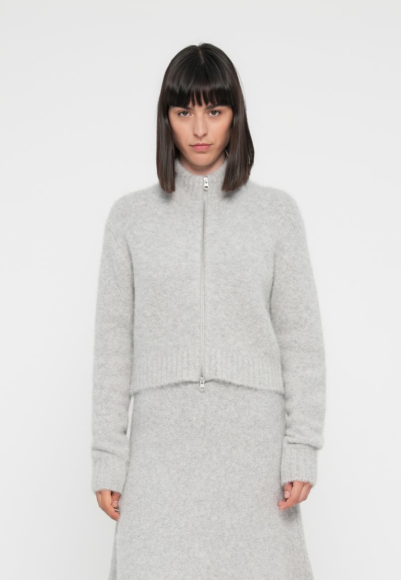 Maglione grigio chiaro con zip, realizzato in materiale morbido e peloso, con colletto alto e orlo a costine. Presenta maniche lunghe e leggermente larghe.