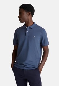 Mladý muž s tmavou pletí a krátkými kudrnatými vlasy, oblečený v modré polokošili a námořnických kalhotách, stojící s rukama v kapsách a dívající se nalevo.