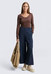 Braunes Langarmshirt, hoch taillierte marineblaue weite Hose, beige klobige Stiefel und eine große beige Tragetasche. Einfaches, klares Design.