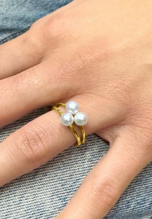 Main sur tissu en denim portant une bague en or avec trois perles blanches disposées en un triangle sur le doigt.