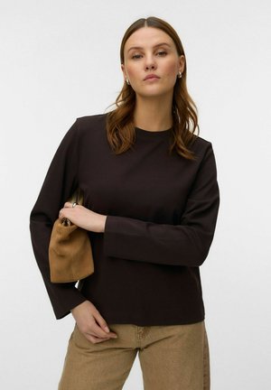 Kvinde med lyst brunt hår iført en sort langærmet top og beige bukser, der holder en lille brun håndtaske under armen.