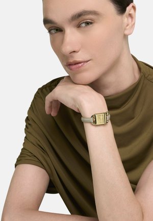 Nahaufnahme einer Person mit heller Haut und grünen Augen, die das Kinn auf die Hand stützt, eine olivgrüne Oberbekleidung und eine goldene Uhr mit beigem Armband trägt.