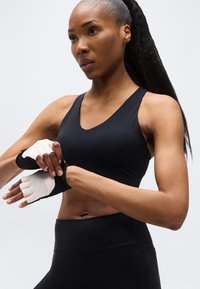 Αθλητική γυναίκα με μαύρα αθλητικά ρούχα που προσαρμόζει λευκά και μαύρα γάντια χωρίς δάχτυλα, προετοιμάζοντας για προπόνηση σε απλό φωτεινό φόντο.