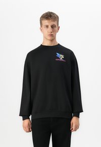 Ung man i en svart sweatshirt med färgglogg O'Neill-logotyp på bröstet, stående mot en enkel vit bakgrund.