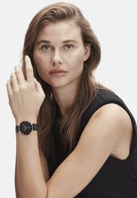 Schwarze Uhr mit rundem Zifferblatt, goldenen Akzenten und einem schwarzen Armband. Das Modell hat lange Haare und stützt ihren Arm auf ihrer Schulter.