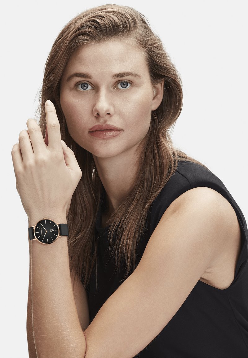 Schwarze Uhr mit rundem Zifferblatt, goldenen Akzenten und einem schwarzen Armband. Das Modell hat lange Haare und stützt ihren Arm auf ihrer Schulter.