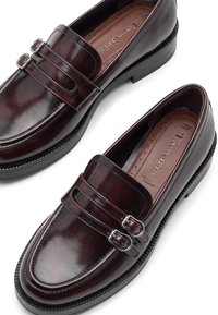 Een paar gepolijste donker bordeauxrode leren loafers met dubbele gespbandjes en zwarte rubberzolen, met bruine gevoerde binnenzolen.