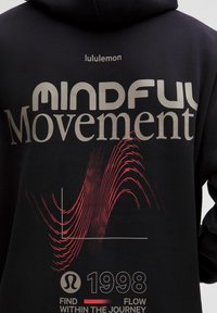 Rückseite eines schwarzen Hoodies mit dem Text "lululemon", "Achtsame Bewegung", rotem Wellendiagramm, lululemon-Logo, "1998" und "Finde den Fluss auf deiner Reise."