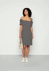 Mulher com cabelo encaracolado, vestindo um vestido preto sem ombros com estampa floral branca, tênis brancos, segurando uma bolsa bege, de pé em frente a uma parede lisa.