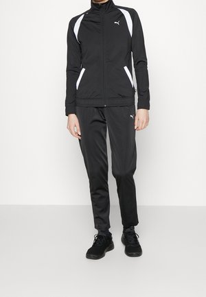 Person trägt einen schwarzen Puma-Trainingsanzug mit weißen Akzenten und schwarze Turnschuhe, steht vor einem schlichten weißen Hintergrund.