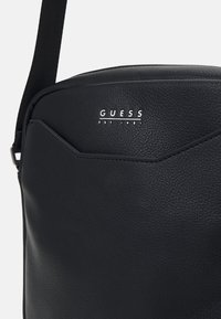 Borsa a tracolla nera in finta pelle testurizzata, con un logo argentato sulla parte anteriore e un design semplice e moderno con una tracolla regolabile.