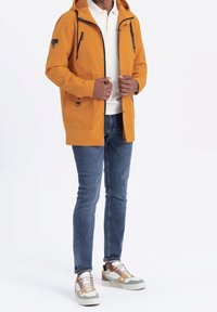 Veste imperméable jaune avec capuche, fermeture éclair à l'avant et plusieurs poches. Portée avec un jean bleu et des baskets multicolores.