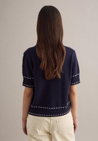 Mujer con cabello largo y castaño, vestida con un suéter de manga corta azul marino con ribetes blancos y pantalones beige claros, de espaldas frente a una pared lisa.