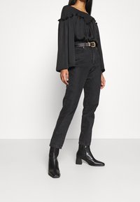 Mujer lleva una blusa negra de manga larga con volantes metida en unos pantalones vaqueros negros de cintura alta, con un cinturón con tachuelas y botas de tobillo negras de tacón.