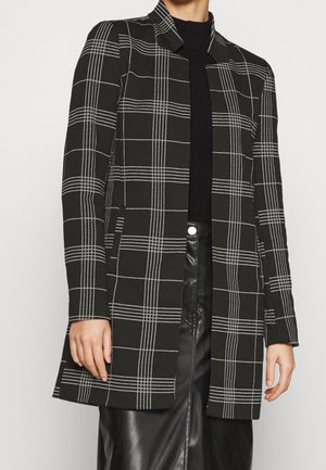 Abrigo largo de cuadros blancos y negros con corte recto, que presenta un cuello de muesca y sin cierres visibles, llevado sobre un suéter oscuro.