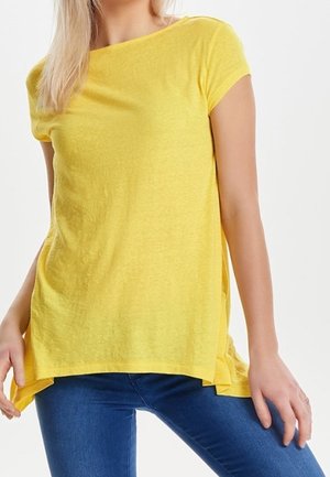 Vrouw draagt een losse gele top met korte mouwen gecombineerd met een blauwe spijkerbroek tegen een effen achtergrond.