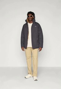 Σκούρο γκρίζο μπουφάν με γούνα και κουκούλα, με φερμουάρ μπροστά και αντίθετη καφέ επένδυση. Συνδυάζεται με μπεζ παντελόνι και λευκά αθλητικά παπούτσια.