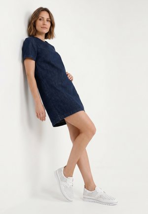 Woman in a short-sleeve dark blue dress and white perforated sneakers, leaning against a white wall with one hand on her hip.