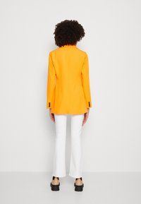 Ljusorange kavaj med slag, skräddarsydd design, i sällskap med vita utsvängda byxor. Svarta platåskor fulländar looken.