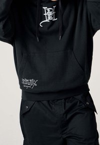 Felpa nera con tasca anteriore e coulisse. Presenta il logo "Don Ed Hardy designs" in bianco sull'angolo in basso a sinistra. Tessuto morbido, vestibilità comoda.