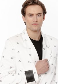 Młody mężczyzna z jasnobrązowymi włosami, noszący białą marynarkę z czarnymi abstrakcyjnymi symbolami na czarnej koszuli, trzymający kołnierz marynarki.