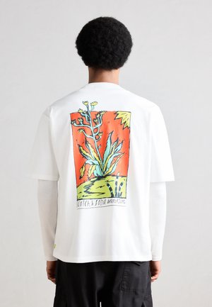 Persona con capelli ricci corti indossa una maglietta bianca con una grafica colorata astratta di piante e sole con il testo "Scotch & Soda Amsterdam" sul retro.