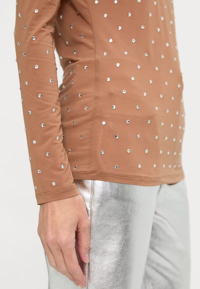 Tricou cu mânecă lungă, de culoare bej, împodobit cu paiete argintii pe întreaga suprafață, asociat cu pantaloni argintii lucioși. Tricoul are un design ajustat și o textură fină.