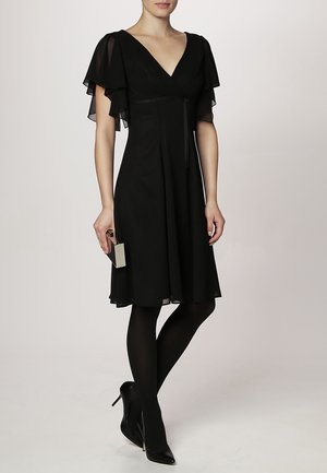 Mujer con vestido negro hasta la rodilla con mangas con vuelo, medias negras y tacones altos negros, sosteniendo un pequeño bolso de mano rectangular.