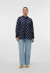 Chemise boutonnée marine à pois blancs, à manches longues, avec col, associée à un jean large bleu clair et des sandales slides jaunes.
